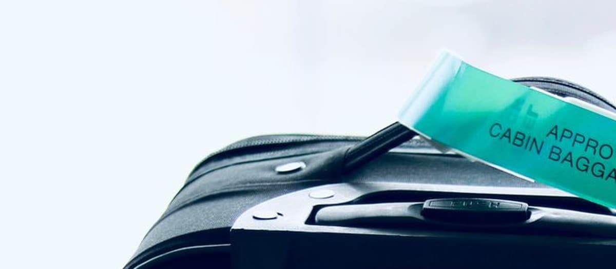The top of a black softside carry-on suitcase with an approved-cabin-baggage tag looped over its handle, against a bright terminal window