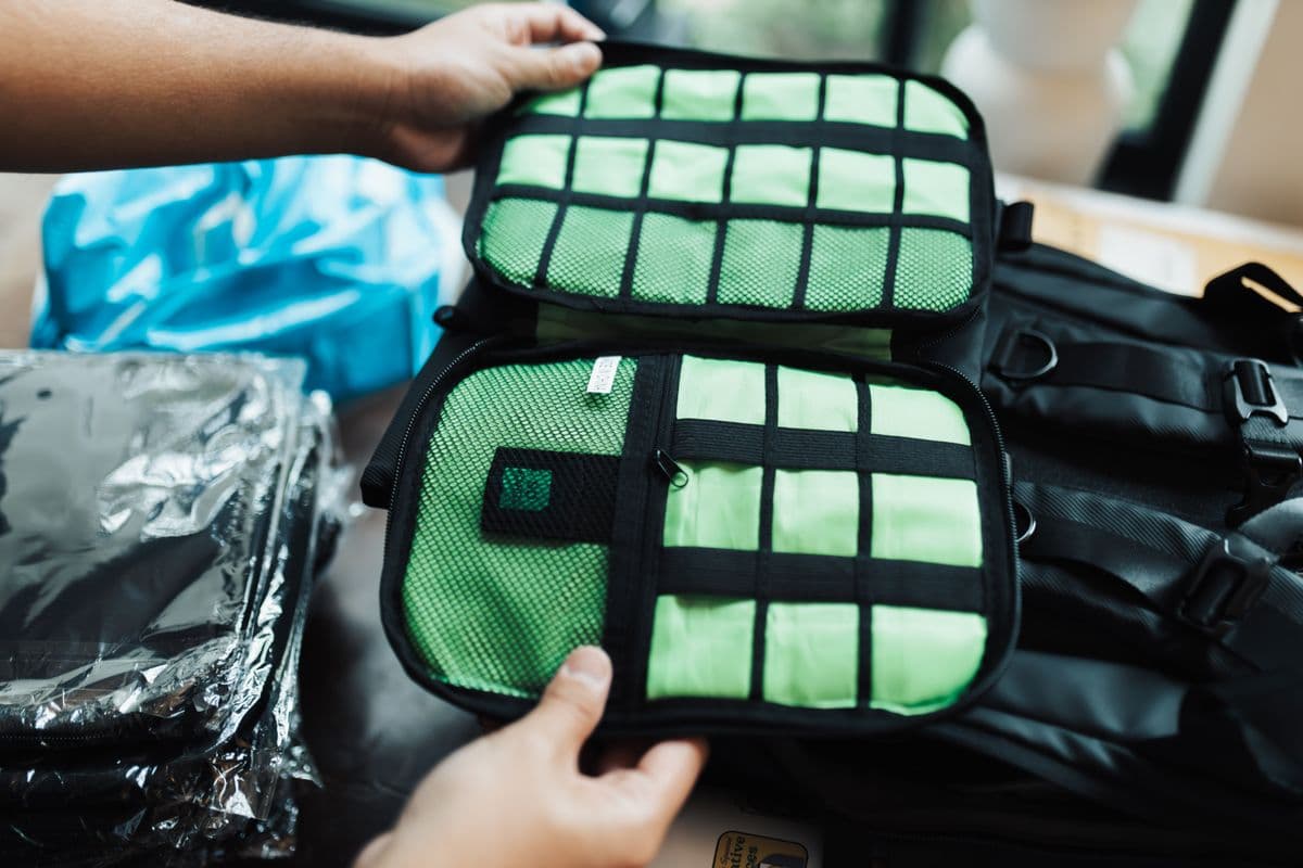 Hands holding open a mesh-topped packing cube filled with folded clothes, beside a travel backpack